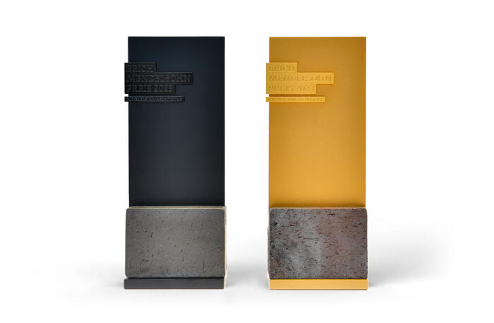 Zwei Siegertrophäen des Erich-Mendelsohn-Preises 2023: links in schwarz, rechts in gold. Beide sind hochkantige Rechtecke, die in einem Backstein-Sockel stehen.
